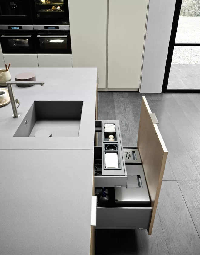 Quadra Collection - Integrated under-sink pull-out storage with grey dividers and a light wood door panel, showcasing architectural functional cleanliness in a modern kitchen.